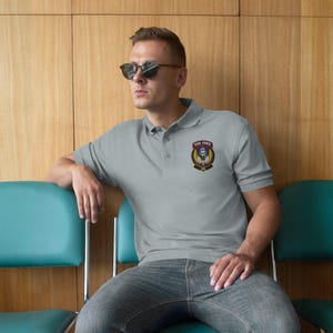 Op de afbeelding: Lichtgrijs poloshirt met een knoopsluiting met drie knopen en korte mouwen. Het shirt heeft een borstpatch met de tekst "TASK FORCE" boven een schedelontwerp en het nummer "141".