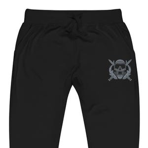 May include: Black sweatpants featuring an elastic waistband with a drawstring. On the right thigh, there is a grey embroidered graphic depicting a scuba diving mask positioned above two crossed diving knives, creating a detailed emblem.