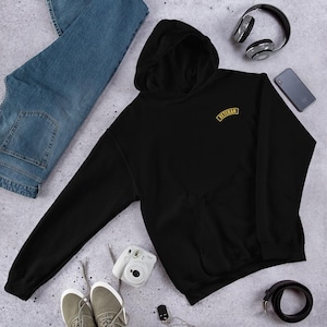 May include: A black hooded sweatshirt with the word "VETERAN" in gold lettering. The sweatshirt is lying on a light gray surface next to a pair of blue jeans, gray sneakers, a camera, headphones, a phone, and a black belt.