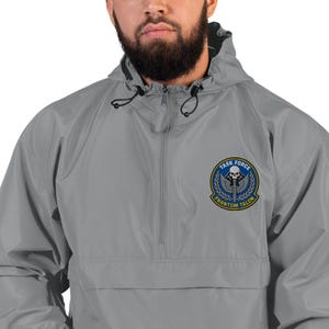 Könnte beinhalten: Graue Kapuzenjacke mit halbem Reißverschluss. Die Jacke hat einen Aufnäher auf der linken Brust mit den Worten "TASK FORCE" über einem Totenkopf- und Schwertdesign und "PHANTOM TALON" darunter.