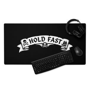 Op de afbeelding: Een zwarte, rechthoekige bureaumat met een witte afbeelding van een scrollbanner met de tekst HOLD FAST, een schedel met gekruiste beenderen en een zeilschip. Een zwart computertoetsenbord, een computermuis en een koptelefoon liggen erop.