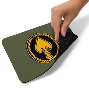 Op de afbeelding: Olijfgroene rechthoekige muismat met een goud en zwart embleem van een speerpunt in een touw cirkel. De muismat heeft een zwarte, getextureerde onderkant.