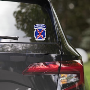 Könnte beinhalten: Ein Aufkleber auf einer Autoscheibe mit dem Abzeichen der 10. Gebirgsdivision. Das Emblem ist ein blauer Schild mit zwei gekreuzten roten Schwertern und einem weißen Reiter oben, der das Wort MOUNTAIN in weißen Großbuchstaben enthält.