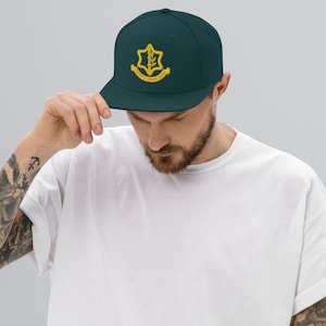 Peut inclure: Casquette de baseball bleu foncé avec une visière plate. La casquette présente un emblème doré avec du texte. La personne sur l'image porte un t-shirt blanc.