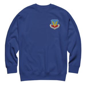 May include: Royal blue sweatshirt with a colourful emblem on the upper left chest. The emblem features a sword, wings, and the words "Air Combat Command". The sweatshirt is made of a soft material and has a crew neck.