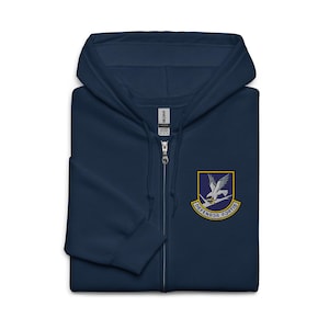 May include: A navy blue zip-up hoodie featuring a hood with drawstrings and a full-length front zipper. On the left chest, there is an embroidered patch depicting a white falcon perched on a branch with the text Defensor Fortis below it.