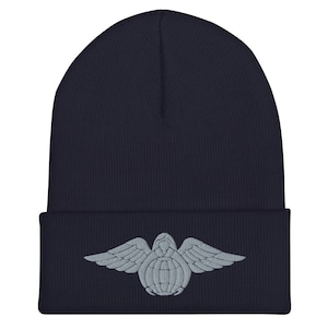Puede incluir: Un gorro azul marino con un diseño bordado gris de una tortuga con alas.