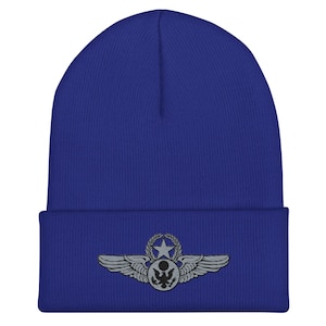 May include: A royal blue beanie hat with a silver emblem featuring wings, a star, and a wreath. The emblem is embroidered on the front of the hat. The hat is made of a soft, knitted material.