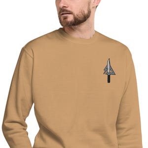 May include: A tan-colored sweatshirt with a crew neck. The sweatshirt features a small embroidered design on the upper left chest, a stylized knife with a lightning bolt and a triangle.
