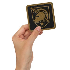 Peut inclure: Écusson carré brodé avec un motif de bouclier orné d'un casque, d'une épée et d'une étoile dorés et noirs. Le patch a une bordure beige et est tenu dans une main.