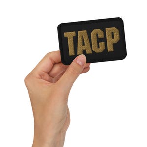May include: Black rectangular patch with the embroidered letters "TACP" in gold. The patch is held in a person's hand against a white background. The patch has a black border.