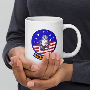 May include: White ceramic coffee mug with a blue, red, and white graphic of a cartoon cat wearing a pilot's uniform and holding a gun. The graphic is surrounded by a blue circle with white stars and the text "TOMCAT" in yellow.