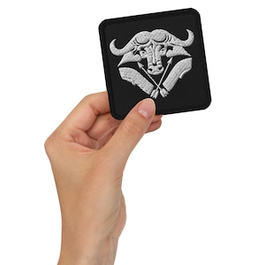 Peut inclure: Un écusson brodé noir avec un motif de tête de buffle blanc. Le buffle a deux flèches qui se croisent devant lui.