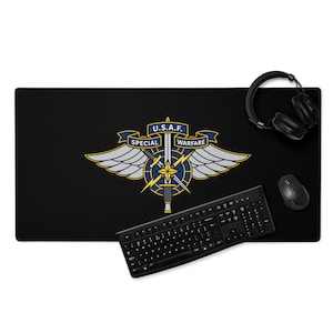 Può includere: Tappetino per scrivania nero con l'emblema U.S.A.F. Special Warfare in blu, oro e argento. L'emblema include ali, una spada e le parole "SPECIAL" e "WARFARE". Sono visibili anche una tastiera, un mouse e delle cuffie neri.