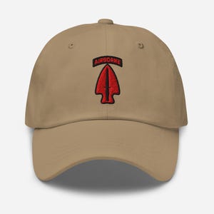 May include: A beige baseball cap with a curved peak. The cap has a red arrow emblem with the word "AIRBORNE" embroidered above it in black. The cap is made of a soft material and has a classic six-panel design.