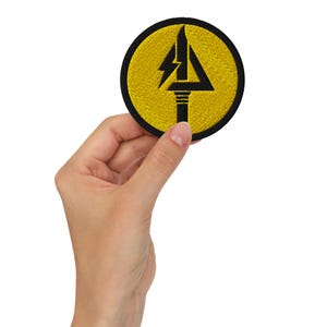 May include: Embroidered patch with a yellow circular background and a black border. The patch features a black design of a lightning bolt above a stylized arrow and a pillar. The patch is held in a hand.