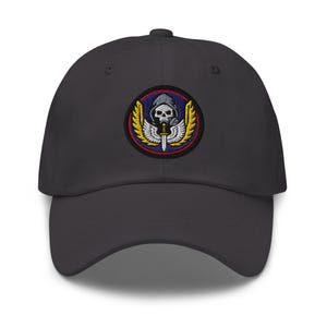 Op de afbeelding: Een donkergrijze baseballcap met een geborduurde patch. De patch toont een schedel met een kap, een zwaard en vleugels, in rood, geel en blauw. De cap heeft een gebogen klep en een verstelbare stoffen band.