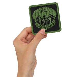 Könnte beinhalten: Ein gestickter Aufnäher in Grün und Schwarz mit dem Text "JTAC" und "THAT OTHERS MAY DIE". Der Aufnäher zeigt eine stilisierte Fledermaus mit einer Kugel in ihren Krallen.