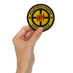 May include: A round embroidered patch with the words "KILLSTREAK MASTER UNTOUCHABLE" in yellow lettering on a black background. The center features a yellow target design with red vertical lines. The patch is held by a hand.