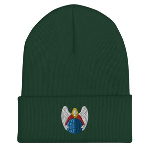 Puede incluir: Un gorro de lana verde oscuro con un ángel bordado en blanco que sostiene un globo terráqueo azul. El ángel lleva una túnica roja y tiene el pelo amarillo.