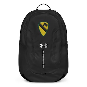 May include: Black Under Armour backpack with a yellow and black emblem. The backpack has a top handle, side pockets, and the Under Armour logo in silver. The backpack is designed with a curved shape and a black zipper.