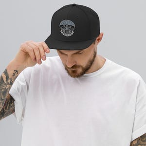 May include: Black baseball cap with a flat brim. The cap features an embroidered design of a parachute, skull, and globe, with the text "THAT OTHERS MAY LIVE" in white. The cap is worn by a person.
