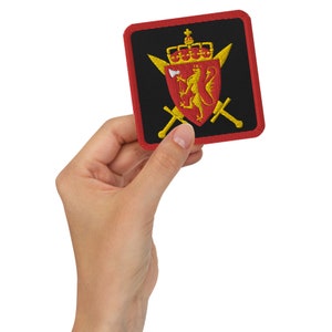 May include: A red, black and gold embroidered patch featuring a lion, crown and crossed swords. The patch is being held in a hand.