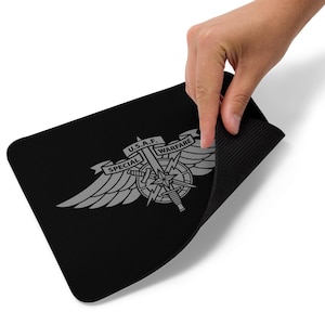 Könnte beinhalten: Schwarzes rechteckiges Mousepad mit silbernem Emblem. Das Emblem trägt die Aufschrift "U.S.A.F. SPECIAL WARFARE" und zeigt Flügel, einen Kompass, ein Schwert und einen Blitz. Eine Hand hebt eine Ecke des Mauspads an.