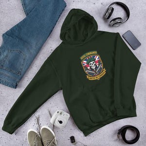 Op de afbeelding: Donkergroene hoodie met een voorzak en een capuchon. De hoodie heeft een patch met de tekst "ROYAL COMMANDO" en "UNITED WE CONQUER" met een schedel- en gekruiste messen-ontwerp. De hoodie wordt gecombineerd met een spijkerbroek, een koptelefoon en een smartphone.