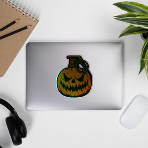 May include: A sticker depicting a pumpkin-shaped grenade with a carved jack-o'-lantern face. The design uses a gradient of orange, yellow, and green, with a black outline. The top of the grenade is dark brown with a black ring.