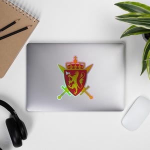 May include: A holographic sticker displaying a red and yellow coat of arms, including a crown, lion, axe, and crossed swords. The sticker is affixed to a laptop. The design is likely related to heraldry or a national symbol.