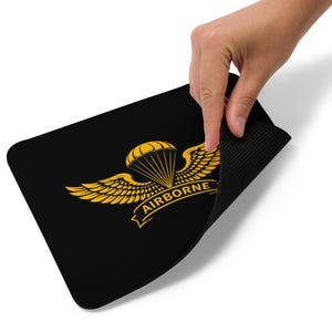 Könnte beinhalten: Schwarzes rechteckiges Mousepad mit goldenem Airborne-Insignien. Das Insignien zeigt einen Fallschirm, Flügel und das Wort "AIRBORNE". Eine Hand hebt das Mousepad an und zeigt die strukturierte Unterseite.