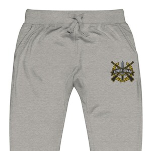 May include: Light gray sweatpants featuring an embroidered emblem on the right leg. The emblem displays two crossed rifles behind a central sword, all encircled by a gold laurel wreath. A black banner across the front reads HONOR GUARD.