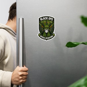 May include: A rectangular magnet with a military-themed design. The magnet features a skull wearing a helmet, crossed rifles, and wings, with the words "BLACK OPS" and "SPECIAL OPERATIONS" in white text on a green and camouflage background.