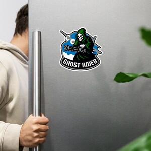 Puede incluir: Una pegatina con un personaje de la Parca verde con una calavera blanca, sosteniendo una ametralladora. El personaje está sobre un fondo azul y blanco con las palabras "GHOST RIDER" en una fuente curva.