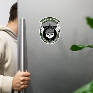 Może przedstawiać: Okrągły magnes z czaszką w berecie i spadochronem. Magnes ma napisy "SPECIAL WARFARE" i "SPECIAL RECONNAISSANCE" w zielono-czarnej kolorystyce. Magnes jest przymocowany do srebrnej lodówki.
