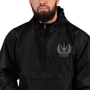 Peut inclure: Veste noire à capuche avec fermeture éclair partielle. L'écusson gris affiche un crâne, des ailes et le texte "JTAC". Le tissu semble lisse et potentiellement imperméable.