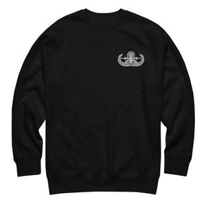 May include: A black long-sleeve sweatshirt featuring an embroidered silver-toned EOD badge on the upper left chest. The badge depicts a bomb emblem centered between two lightning bolts, all surrounded by a laurel wreath design on a dark background.