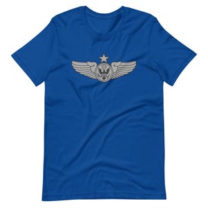 May include: A royal blue t-shirt featuring a silver emblem with wings, a star, and a circular design with an eagle. The short-sleeved shirt is made of a soft material and is suitable for casual wear.