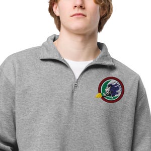 Peut inclure: Un sweat-shirt gris à fermeture éclair quart de zip avec un sous-vêtement blanc. Le sweat-shirt présente un écusson brodé circulaire avec un crâne portant une coiffe à plumes, entouré de vert et de rouge, avec une flamme jaune.