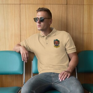 May include: A pale beige polo shirt with a collared neckline and short sleeves. The shirt has an embroidered emblem with a jack-o'-lantern design and the text "JACK O' FRAG". The person is wearing sunglasses and sitting on a teal chair.