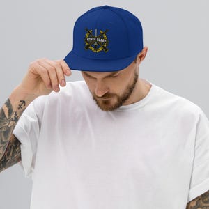 May include: A royal blue snapback cap featuring an embroidered emblem on the front. The design includes two crossed rifles behind a central sword, all framed by a yellow laurel wreath. A banner across the center displays the text HONOR GUARD.