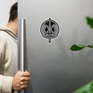 以下が含まれることがあります： 円形エンブレムの中に「SOF TACP」の文字が入った、グレーと黒のステッカー。エンブレムには翼、星、稲妻、垂直の刃が含まれています。ステッカーは銀色の冷蔵庫に貼られています。