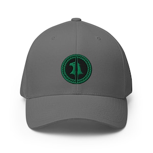 Op de afbeelding: Een grijze baseballcap met een gebogen klep. De cap heeft een cirkelvormige geborduurde patch met een groen ontwerp van een gestileerde adelaar en het nummer "1". Het ontwerp is geplaatst tegen een zwarte achtergrond.