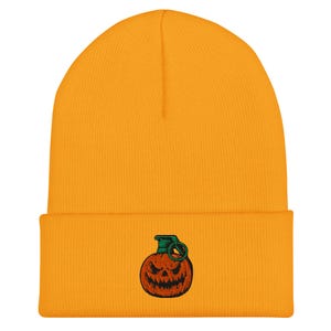 May include: Mustard yellow beanie hat with a turned-up brim. It has an embroidered design of a jack-o'-lantern grenade with a green pin and a scary face. The hat is made of a soft, knitted material.