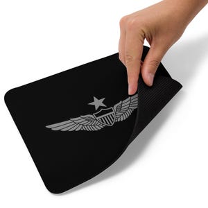Può includere: Tappetino per mouse rettangolare nero con un emblema argentato di ali, una stella e uno scudo. Una mano solleva il bordo, rivelando un lato inferiore strutturato. Il design è un classico simbolo dell'aviazione.