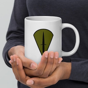 May include: White ceramic mug featuring a black dagger design within a green shield shape. The mug has a standard handle and is held by a person. The design is centered on the mug's surface.