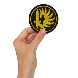 May include: A circular embroidered patch featuring a black background with a vibrant yellow design. The emblem displays a vertical sword positioned next to a stylized wing, all enclosed within a thick yellow circular border with stitched edges.