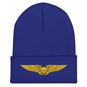 May include: A royal blue beanie hat with a gold-colored emblem featuring a shield and wings. The hat is made of a soft, knitted material and has a folded brim. The emblem is centered on the front of the hat.