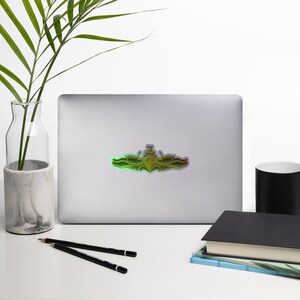 May include: A gold-colored sticker with a submarine emblem. The emblem features a stylized submarine with a diving helmet on top. The sticker has a holographic finish, creating a rainbow effect.